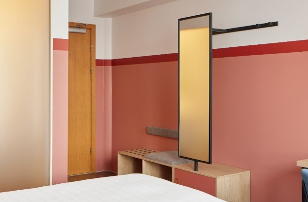 A view of the entrance area of the Standard Plus Room at Center Hotels Plaza in Reykjavík, featuring warm terracotta-colored walls, a wooden bench with cushion, a modern vertical mirror, and