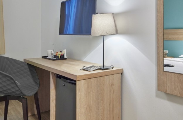A compact and modern workspace in the Standard Single Room at Center Hotels Plaza in Reykjavík, featuring a wooden desk with a reading lamp, wall-mounted TV, tea station, and a reflection of