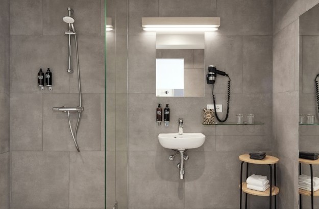 The bathroom of the superior room at Center Hotels Plaza with gray tiles on floor and walls, walk-in shower, sink, mirror, hairdryer and soap dispensers.