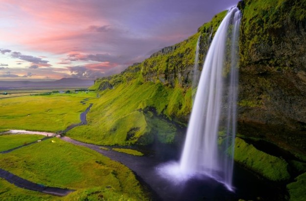 A scenic view of Seljalandsfoss waterfall in Iceland at sunset, surrounded by lush green fields and walking paths, representing Center Hotels’ commitment to sustainability through Green Key e