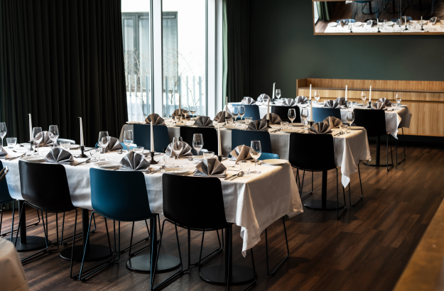 A overview of three tables set up for a venue. With white cloth, cutlery and brown napkins. The light flows into the room from a large window.