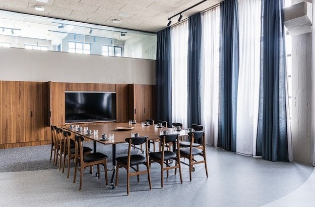 A large meeting room table seen from one side on a floor with glasses on table. A large TV on one of the wall. 