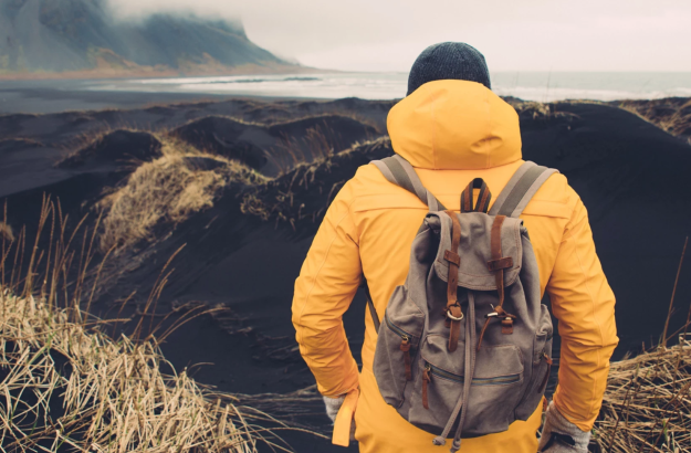 A person standing infront of a scenic landscape in Iceland with the view of mountains and ocean, wearing a yellow jacket, black hat, white gloves and a backpack.
