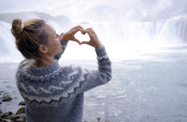 A woman forming a heart with hear hands wearing a lopapeysa sweater in front of Godafoss waterfall in Iceland.
