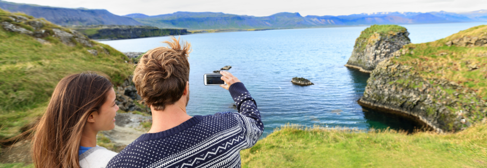 People standing in front of a mountain and lake view in Iceland, taking a picture with a camera dressed in Icelandic wool sweaters.