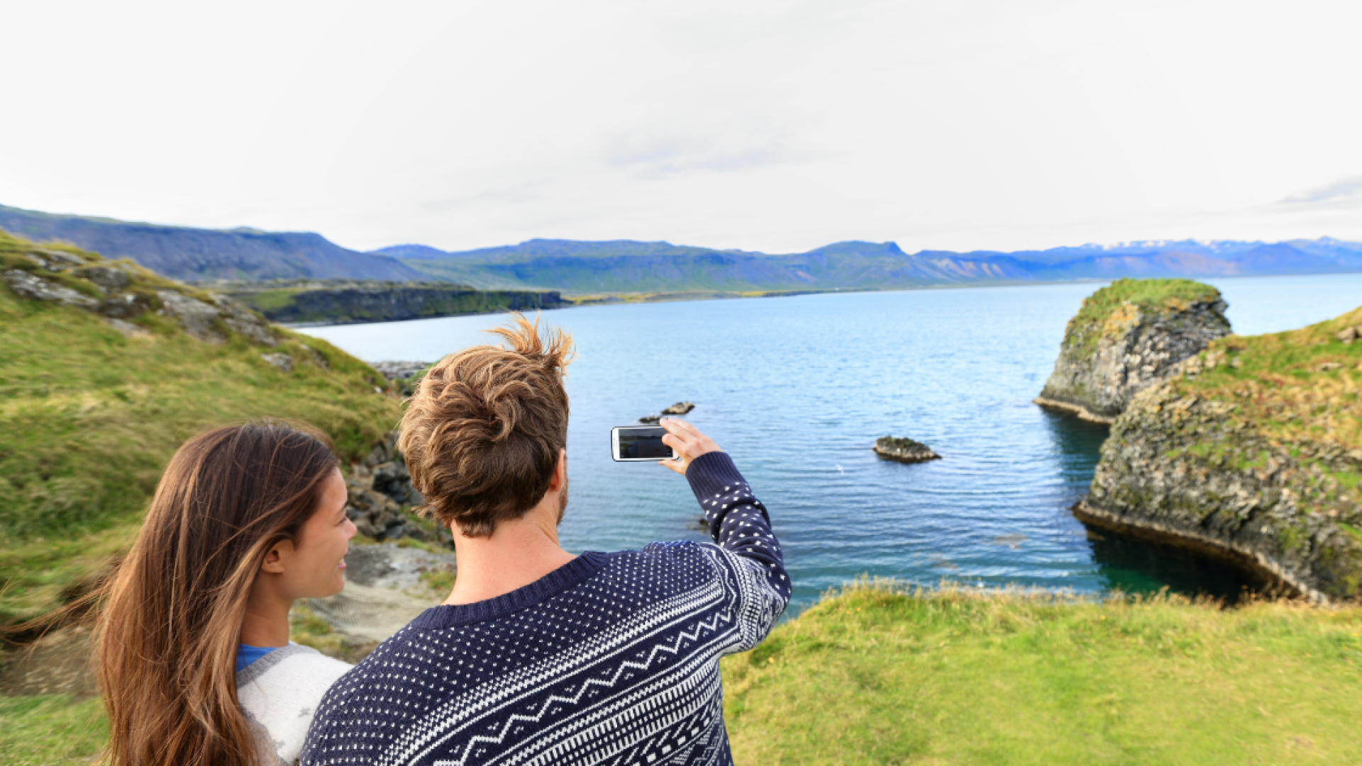 People standing in front of a mountain and lake view in Iceland, taking a picture with a camera dressed in Icelandic wool sweaters.