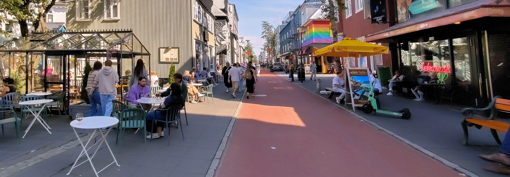 People relaxing at outdoor café tables on Laugavegur in Reykjavík, Iceland, with colorful buildings, shops, restaurants, and pedestrians walking along the busy street on a sunny day.