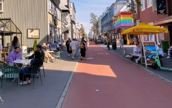 People relaxing at outdoor café tables on Laugavegur in Reykjavík, Iceland, with colorful buildings, shops, restaurants, and pedestrians walking along the busy street on a sunny day.