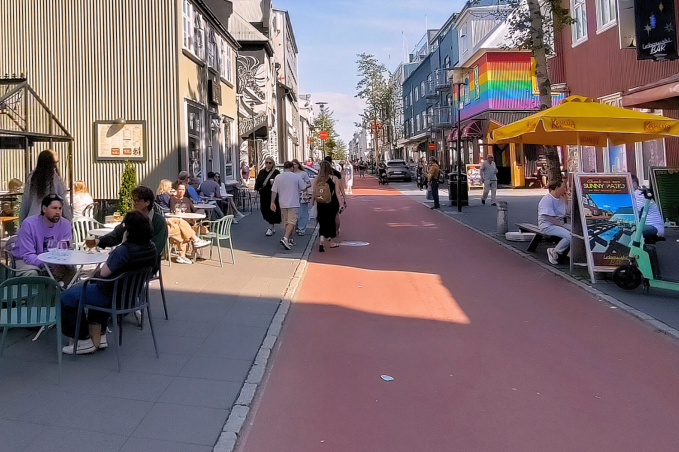 People relaxing at outdoor café tables on Laugavegur in Reykjavík, Iceland, with colorful buildings, shops, restaurants, and pedestrians walking along the busy street on a sunny day.