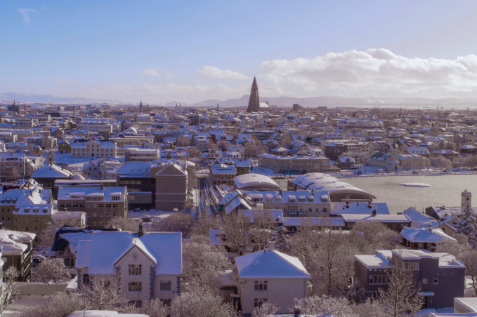 Winter view of downtown Reykjavík with snow-covered rooftops, Hallgrímskirkja church in the center, and leafless trees in the foreground.