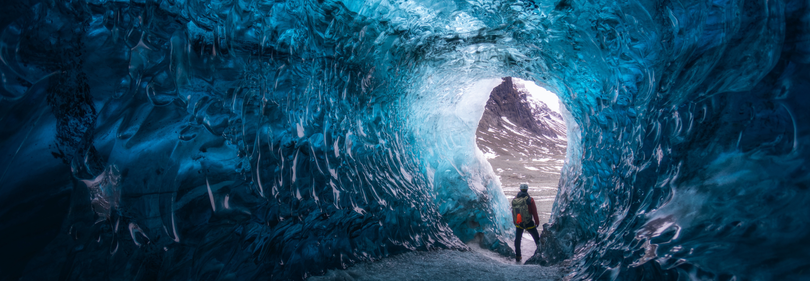 Person standing at the entrance of a bright blue ice cave in Iceland during winter.