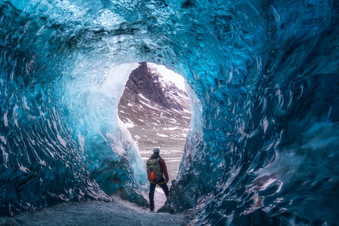 Person standing at the entrance of a bright blue ice cave in Iceland during winter.