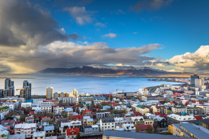 Panoramic view of Reykjavík with colorful houses, Faxaflói Bay, and snow-covered Mount Esja under a dramatic cloudy sky.