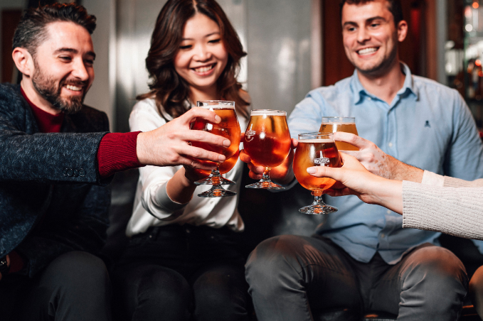 Group of friends smiling and clinking glasses of craft beer while socializing in a cozy bar setting.