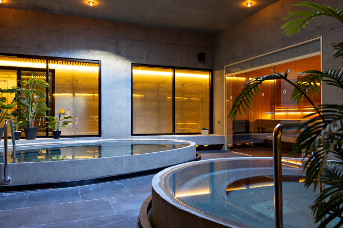 Modern indoor spa area in Reykjavík featuring warm-lit hot tubs, concrete walls, glass sauna, and tropical plants in a relaxing wellness setting.