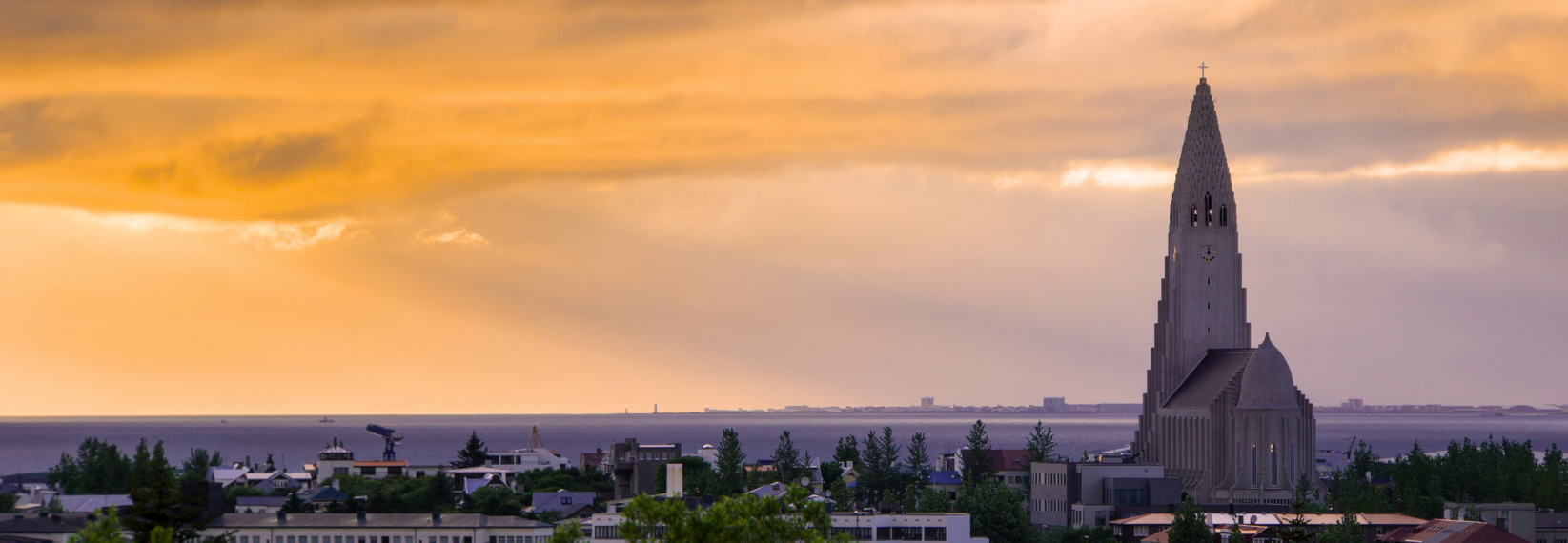 Hallgrímskirkja church rising above Reykjavík skyline at sunset with orange sky and the ocean in the background.