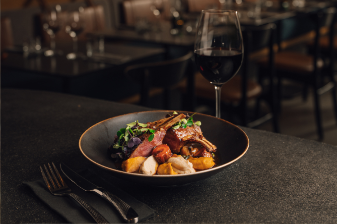 Plated gourmet lamb dish with roasted vegetables and microgreens served on a dark plate with a glass of red wine in a softly lit restaurant.