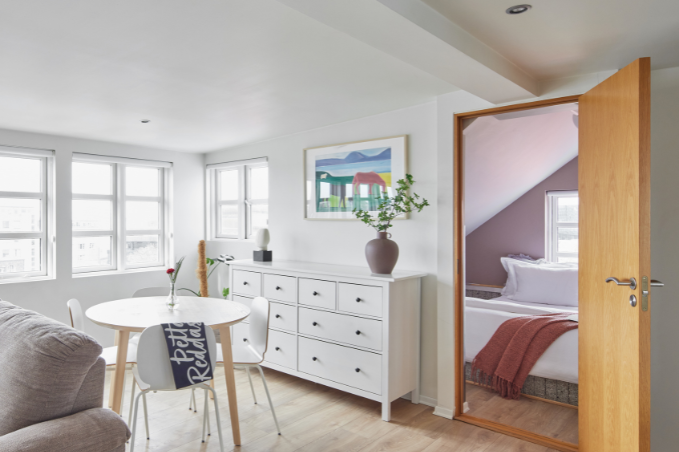 Bright Scandinavian-style apartment interior in Reykjavík featuring a dining table, dresser, large windows, and an open doorway leading to a cozy bedroom.