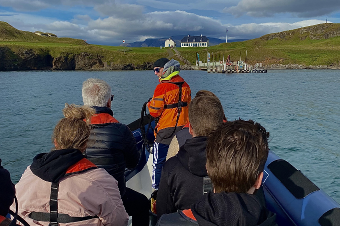 Group of people wearing life jackets on a small boat tour near the Viðey island coastline with green hills and a harbor in the background.