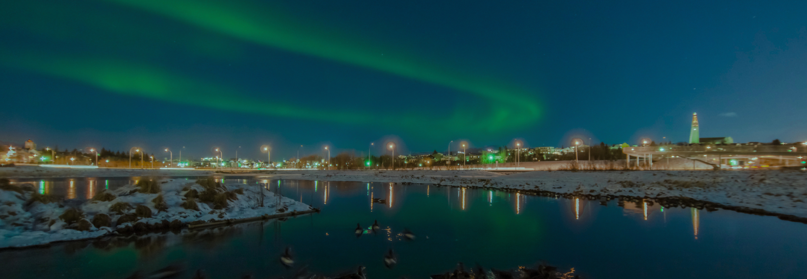 Green northern lights stretching across the sky above Reykjavík with city lights and reflections on a calm, snowy river.