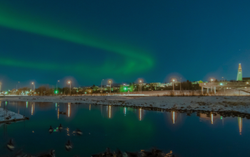 Green northern lights stretching across the sky above Reykjavík with city lights and reflections on a calm, snowy river.