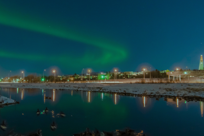 Green northern lights stretching across the sky above Reykjavík with city lights and reflections on a calm, snowy river.