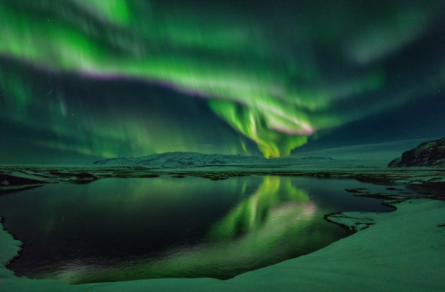 Green and purple northern lights reflecting on a snowy lake in Iceland.