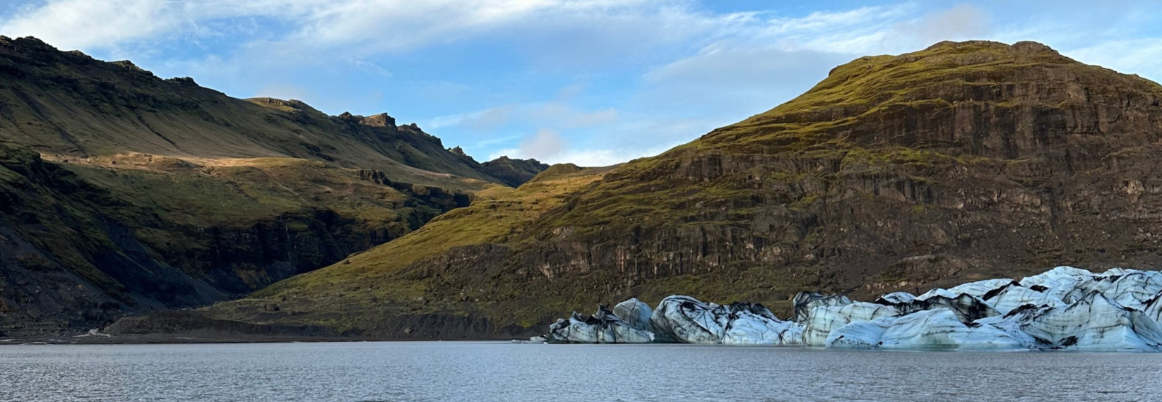 Blue-white glacier ice streaked with volcanic ash beside a calm lagoon, surrounded by rugged Icelandic mountains.