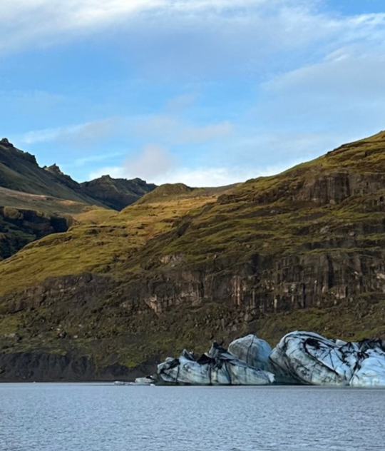 Blue-white glacier ice streaked with volcanic ash beside a calm lagoon, surrounded by rugged Icelandic mountains.