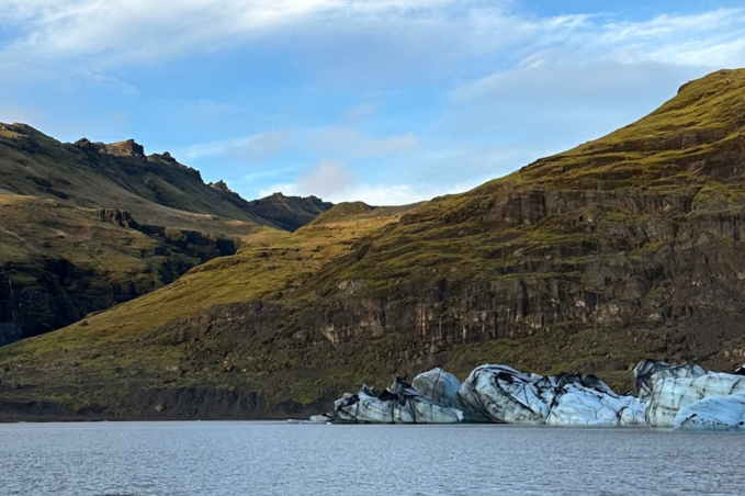 Blue-white glacier ice streaked with volcanic ash beside a calm lagoon, surrounded by rugged Icelandic mountains.