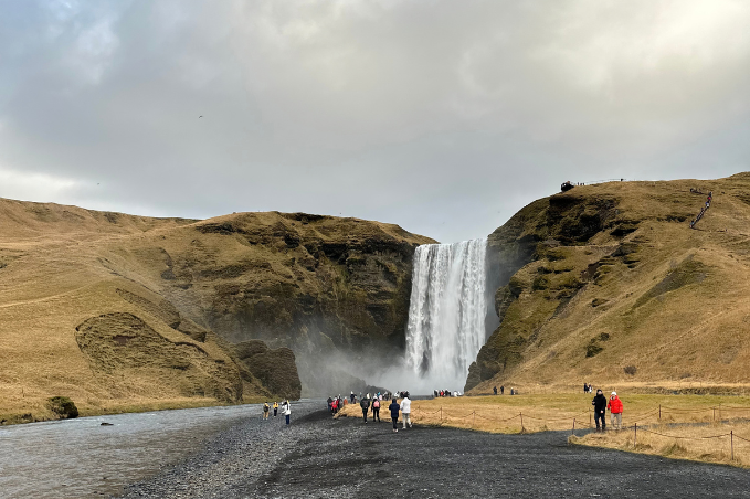 Visitors approaching Skógafoss waterfall along a black gravel path, with grassy cliffs and mist surrounding the cascade.