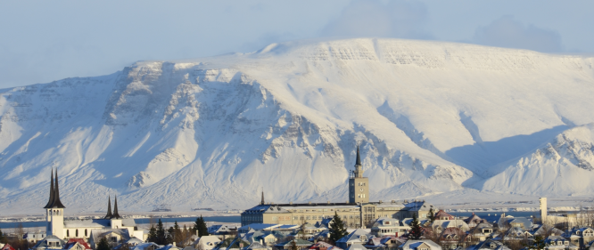 Snowy rooftops in Reykjavik, two dark church spires and a clock-tower building in the foreground, with a massive white mountain ridge behind under a pale blue sky.