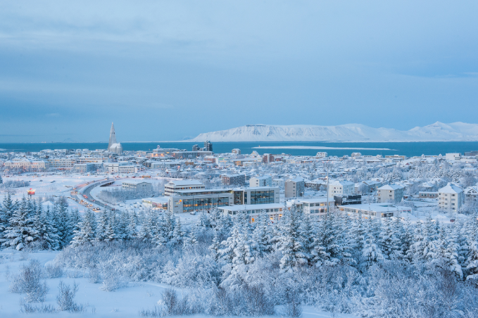 A picture taken over Reykjavik city where everything is fully covered in snow, with Hallgrimskirkja church standing tall in the middle and mountains in the bakcground.