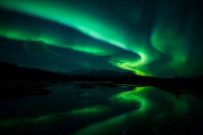 A picture of the green Northern Lights dancing in the sky in Reykjavik Iceland, reflecting in the water beneath.