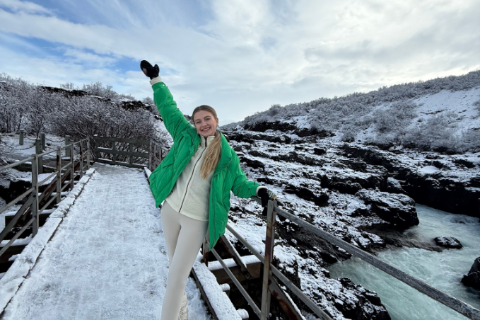 A picture of a girl standing on a snow covered bridge near a river in Iceland wearing a green winter jacket and gloves holding one hand up in the sky.