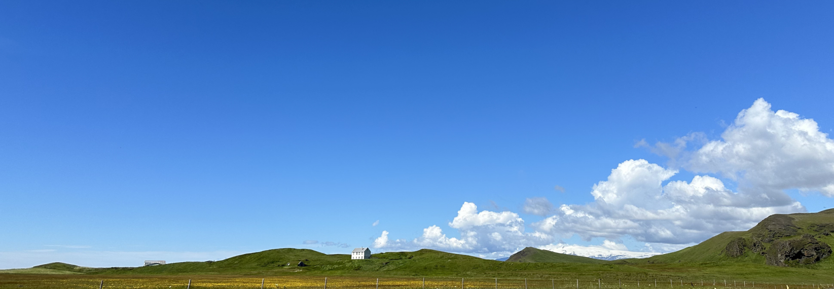 Rolling green hills, a white farmhouse and distant snow-capped mountains beneath a bright blue sky in rural Iceland.