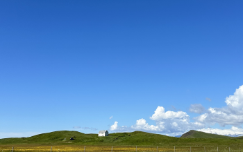 Rolling green hills, a white farmhouse and distant snow-capped mountains beneath a bright blue sky in rural Iceland.