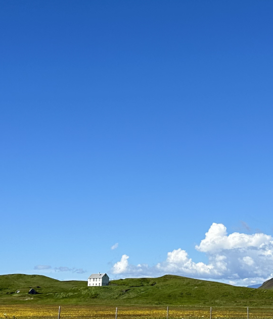 Rolling green hills, a white farmhouse and distant snow-capped mountains beneath a bright blue sky in rural Iceland.