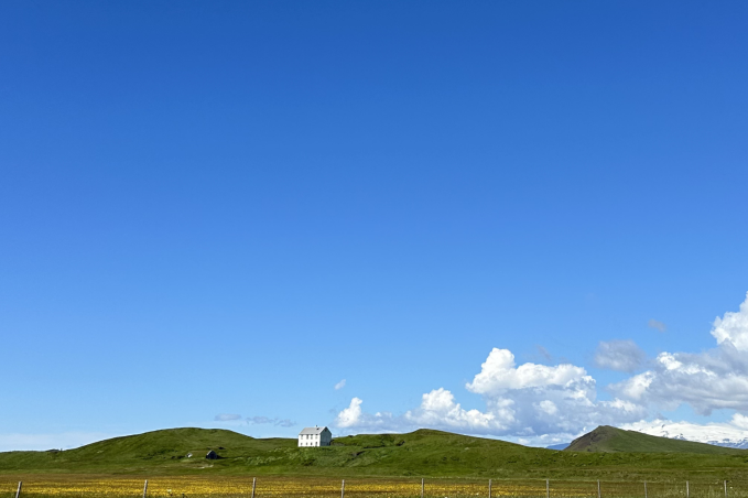 Rolling green hills, a white farmhouse and distant snow-capped mountains beneath a bright blue sky in rural Iceland.