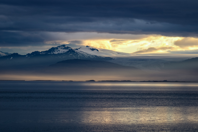 Dramatic view of an Icelandic glacier and mountains beneath dark clouds with golden evening light.