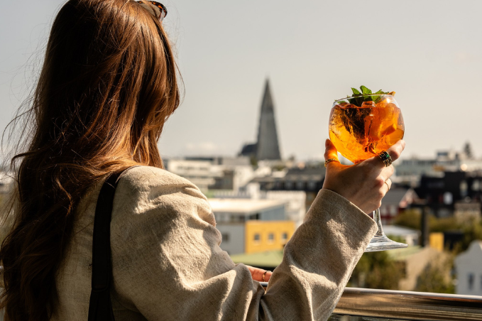 Person holding an orange cocktail on a rooftop terrace overlooking Reykjavík with Hallgrímskirkja in the distance.