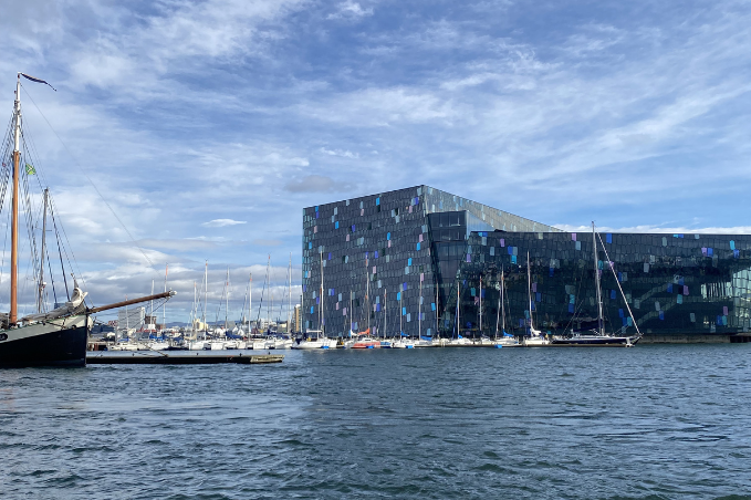 Harpa Concert Hall reflected in Reykjavík Harbour with sailboats moored along the waterfront under a blue sky.