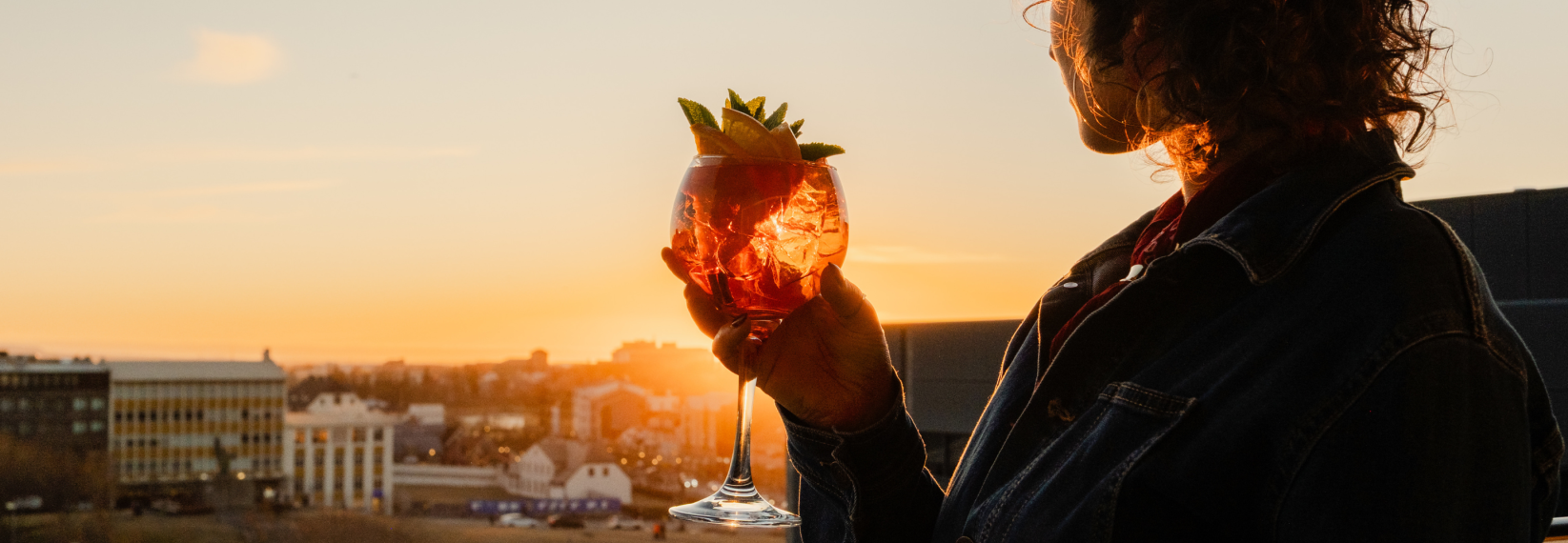 Guest holding a colorful cocktail while enjoying panoramic sunset views over Reykjavík from a rooftop bar.