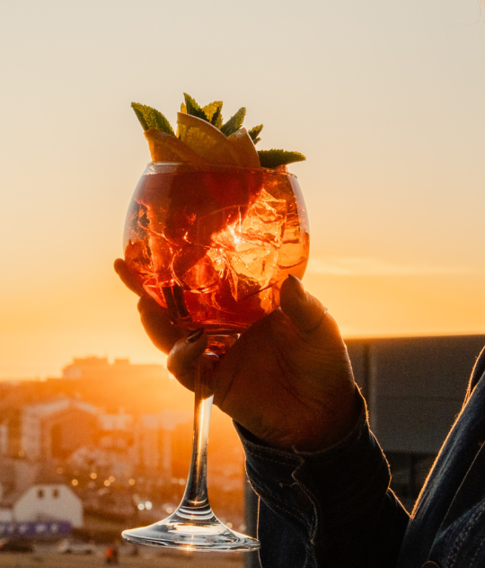 Guest holding a colorful cocktail while enjoying panoramic sunset views over Reykjavík from a rooftop bar.