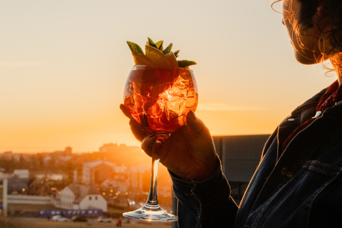 Guest holding a colorful cocktail while enjoying panoramic sunset views over Reykjavík from a rooftop bar.