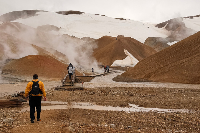 Visitors crossing a wooden bridge through steaming geothermal hot springs surrounded by colorful volcanic hills in Landmannalaugar, Iceland.