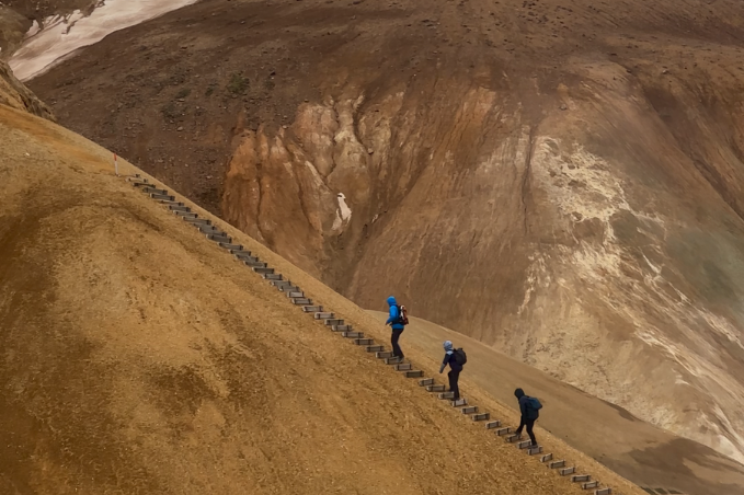 Three hikers climbing the steep wooden staircase on the Brennisteinsalda hiking trail in Landmannalaugar, Iceland.