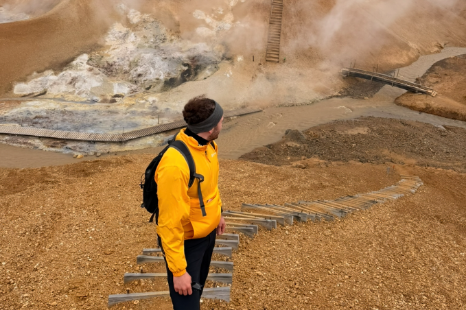 Hiker in a yellow jacket overlooking steaming geothermal vents, colorful volcanic hills, and wooden hiking trails in Landmannalaugar, Iceland.