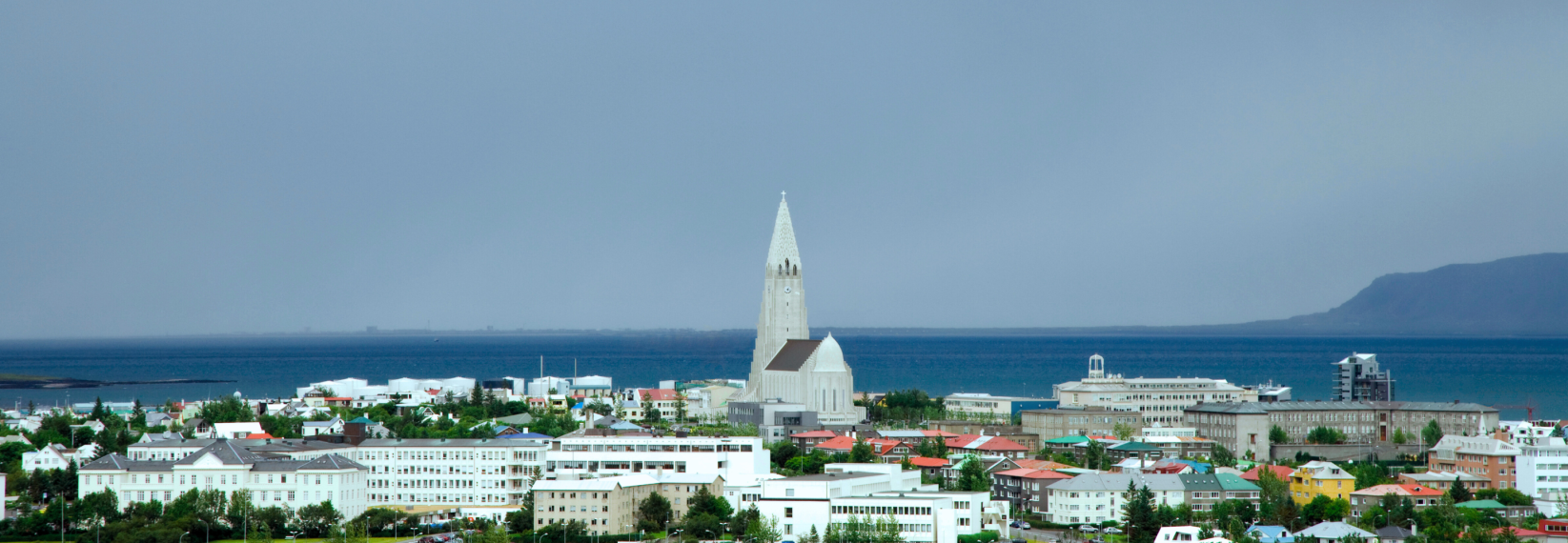 A panoramic view of downtown Reykjavík featuring Hallgrímskirkja, a tall white church with a pointed tower. The city is surrounded by colorful houses and green trees. In the background, the b