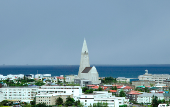 A panoramic view of downtown Reykjavík featuring Hallgrímskirkja, a tall white church with a pointed tower. The city is surrounded by colorful houses and green trees. In the background, the b
